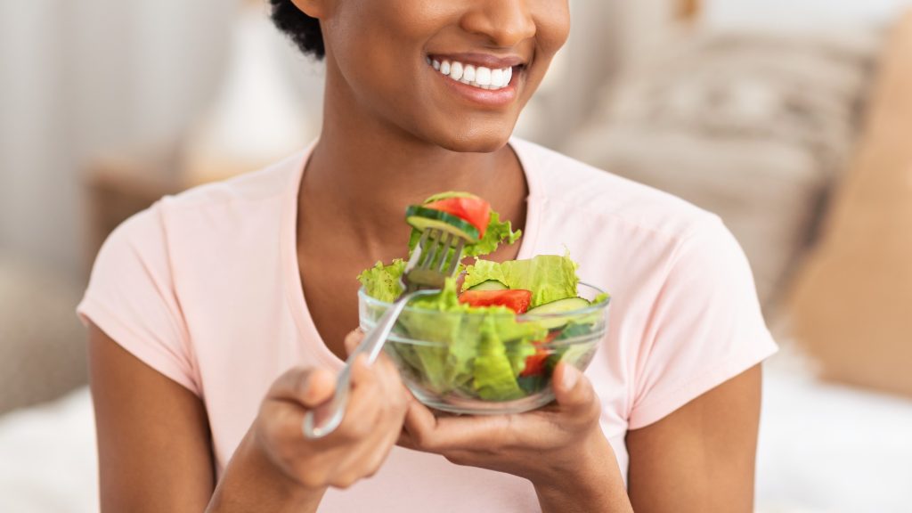 A smiling young Black woman eating a bowl of fresh salad, representing healthy eating habits for gut health.